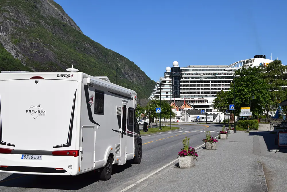 En hvit bobil står parkert i en veikant med potteplanter, en delt vei og et cruiseskip i bakgrunnen under en klarblå himmel. Fjell er synlige i det fjerne, og fanger essensen av Eidfjord – der sport og fritid blomstrer i Norsk natur.