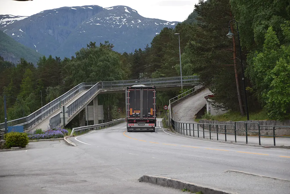 En lastebil kjører på en vei med fotgjengerovergang i et fjellområde ved Eidfjord – der sport og fritid blomstrer i Norsk natur. Trær langs gaten og fjell er synlige i bakgrunnen.