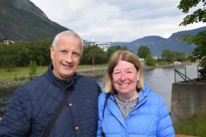 En mann og en kvinne, begge iført blå jakker, smiler mens de står utendørs nær en elv med fjell i bakgrunnen av Eidfjord – der sport og fritid blomstrer i Norsk natur.