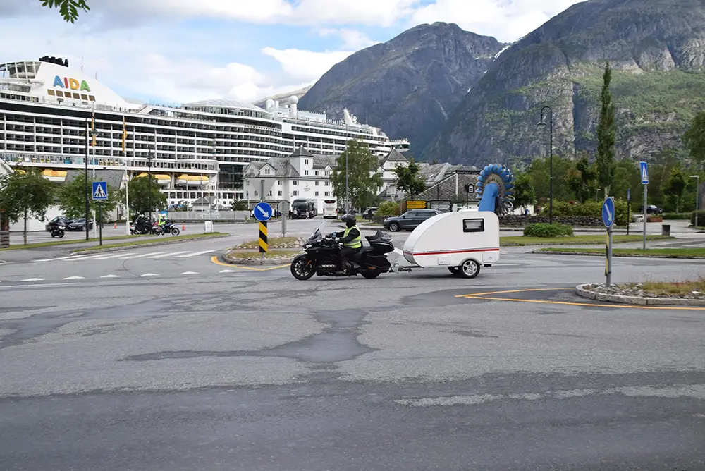 En person kjører motorsykkel og trekker en liten campingvogn i en rundkjøring, med et stort cruiseskip forankret i bakgrunnen og fjellandskapet rundt området – Eidfjord, der sport og rekreasjon trives i norsk natur.