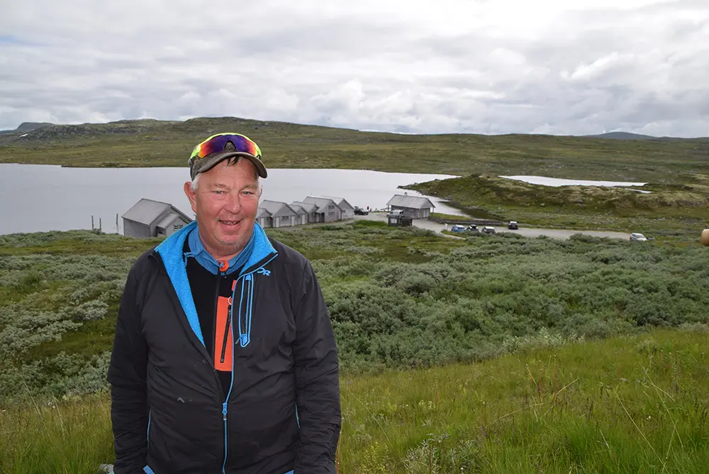 En person i friluftsklær står på en gressbakke med en klynge av små bygninger, en innsjø og bølgende åser i bakgrunnen, og fanger essensen av Eidfjord – der sport og fritid blomstrer i Norsk natur.