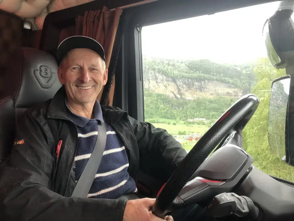 En smilende person med caps og bilbelte sitter i førersetet på et kjøretøy med utsikt over Eidfjord – der sport og fritid blomstrer i Norsk natur, og viser det fantastiske fjellandskapet i bakgrunnen.