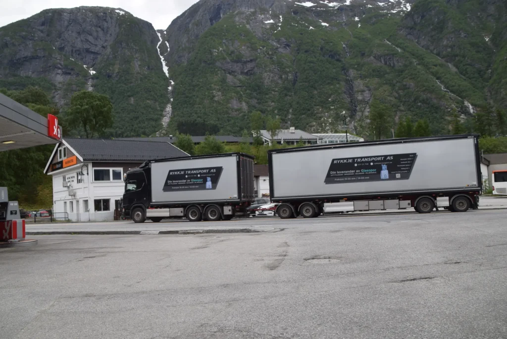 En svart lastebil med en stor tilhenger merket "Rykke Transport AS" står parkert i nærheten av en hvit bygning, satt mot Eidfjordens fantastiske fjellbakgrunn – der sport og fritid blomstrer i Norsk natur.